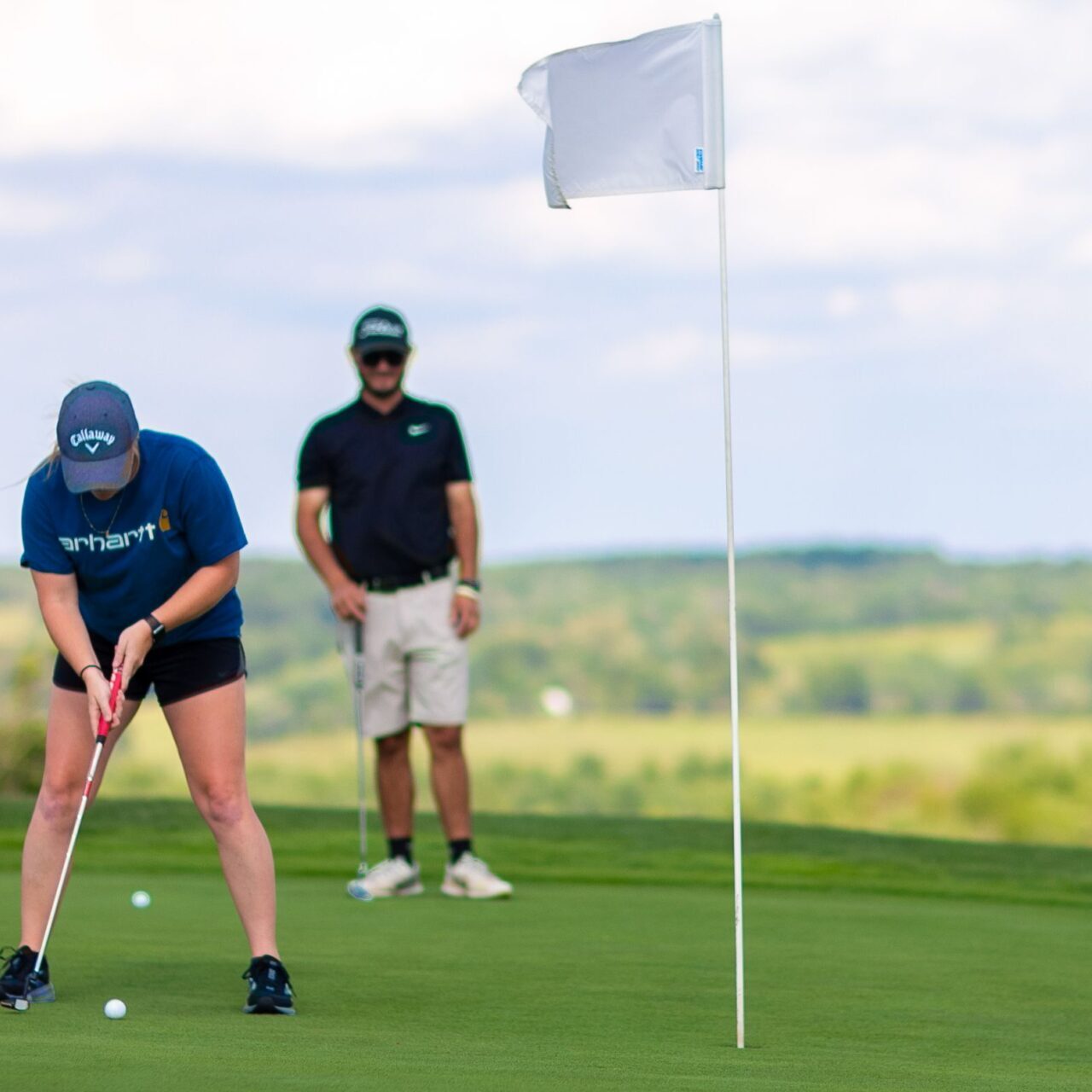 green and spacious golf course with woman putting at hole and man waiting in back with golf club